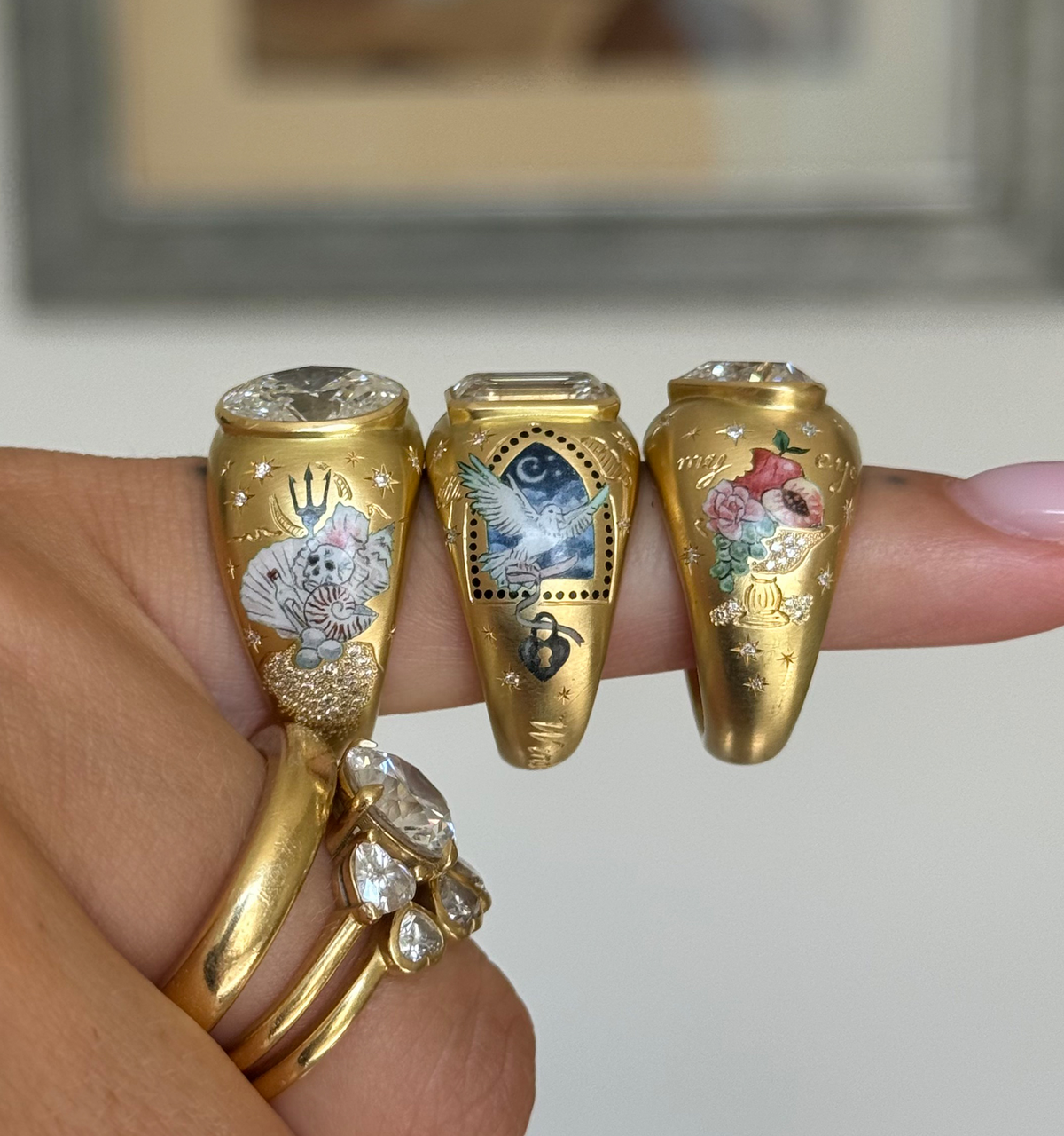 Woman wearing three gold rings with intricate hand-painted designs and a gold band with multiple diamonds.