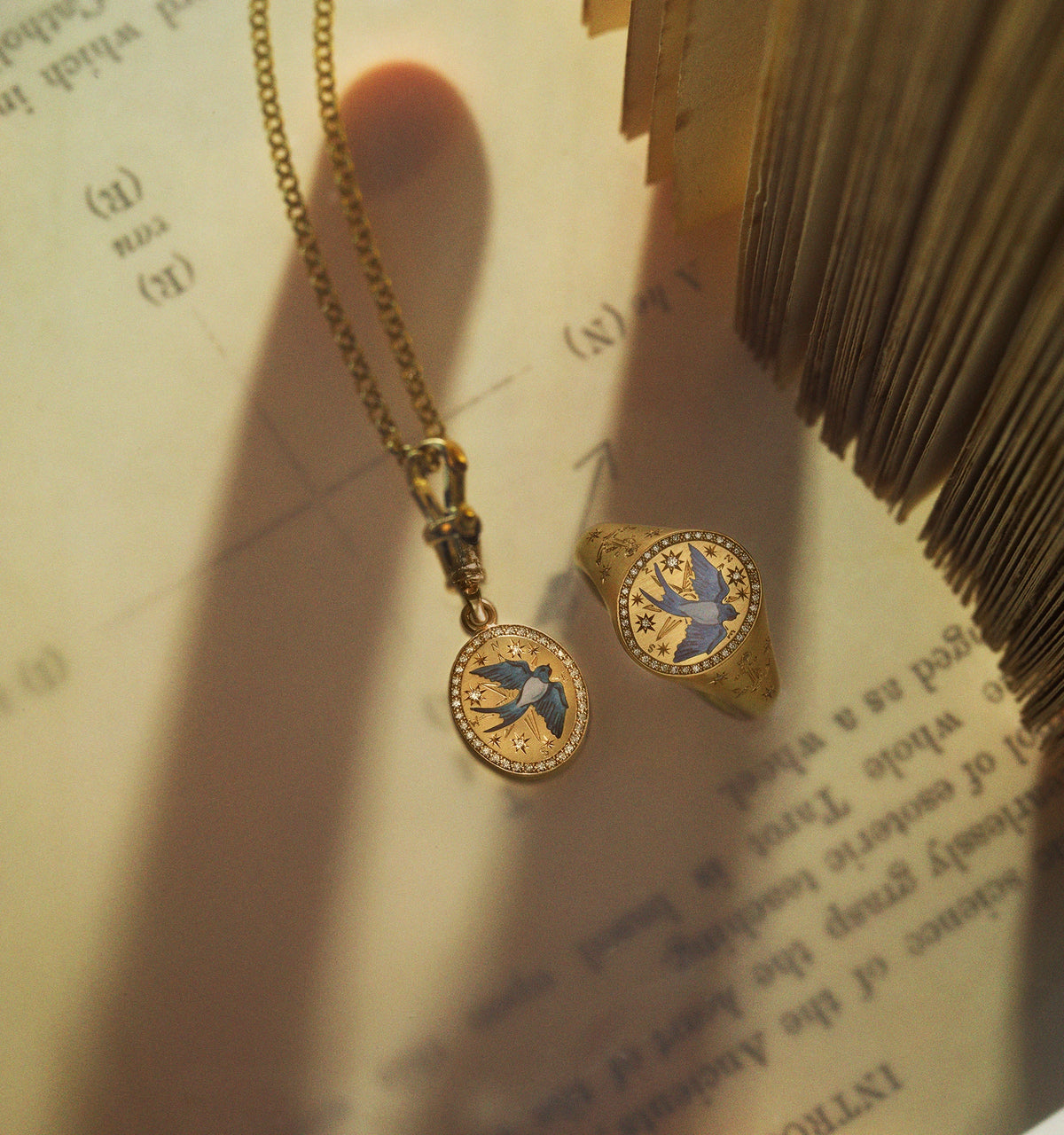 Gold pendant featuring a bird in enamel and gold ring with a similar design on a textured surface.