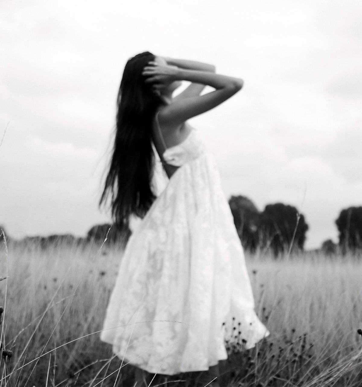 Woman wearing a white dress standing in a field with long grass, hair flowing as she poses against a cloudy sky.