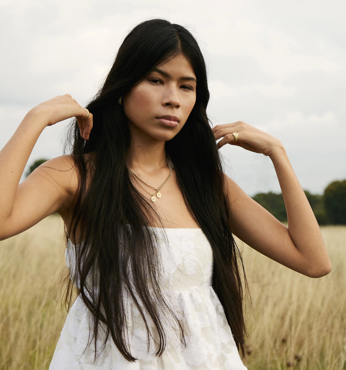 Woman wearing layered gold necklaces and a gold ring, standing in a field with long grass and a cloudy sky.