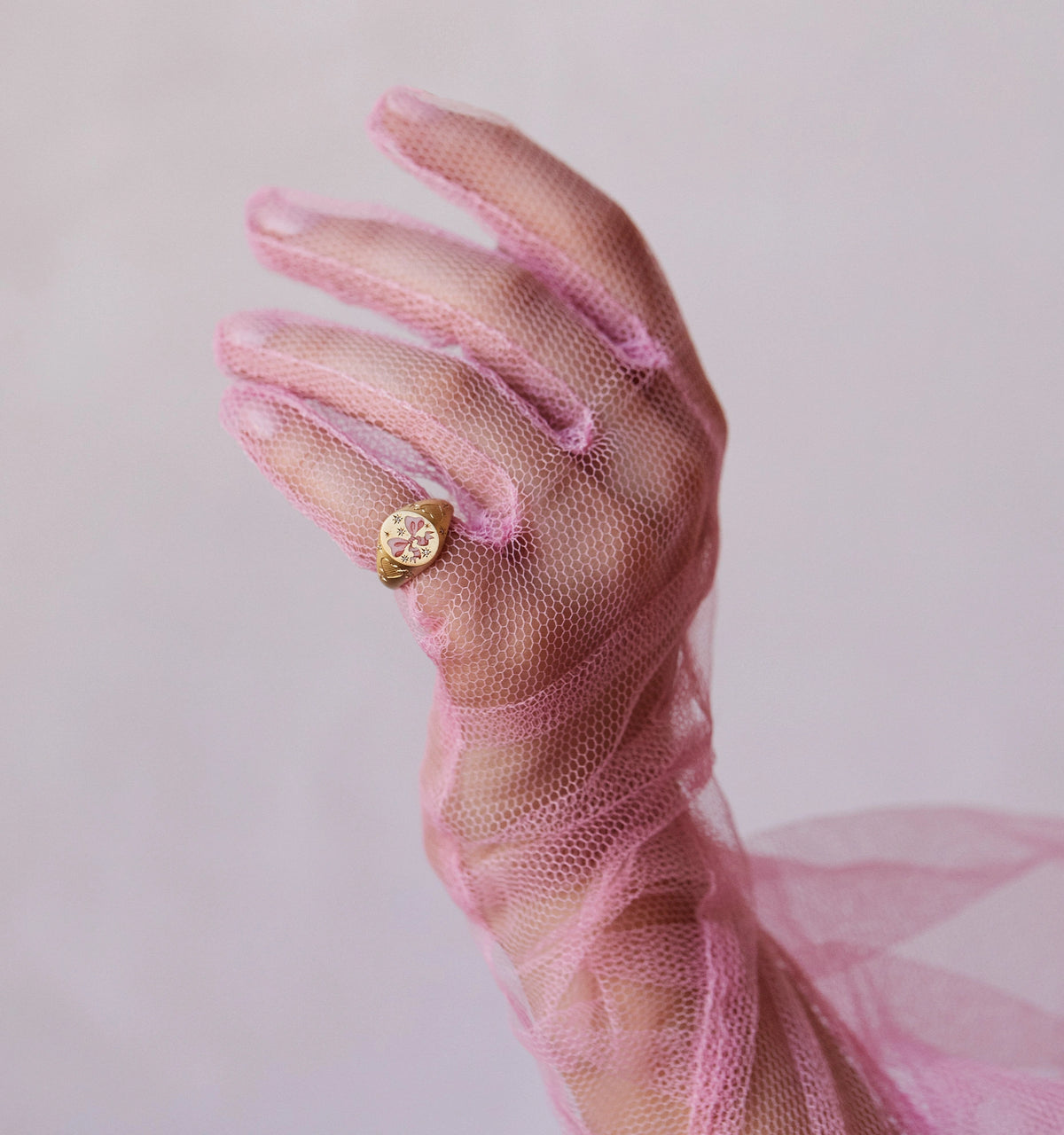 Woman wearing a gold ring with an engraved floral design, hand covered in sheer pink fabric.