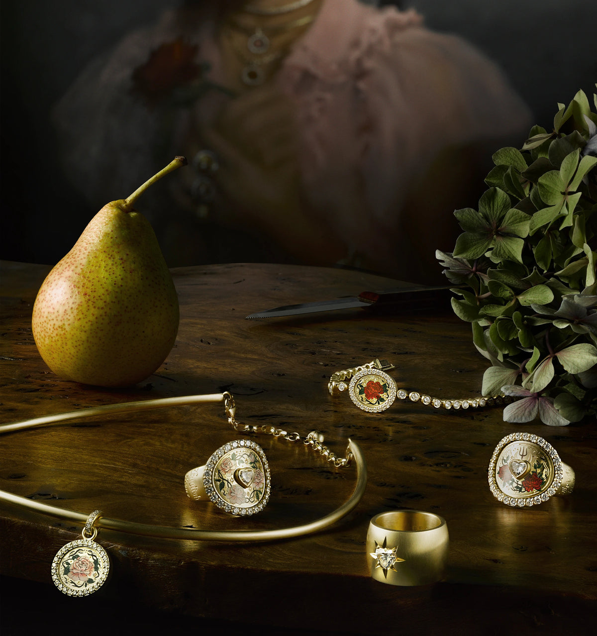 Gold jewellery pieces arranged on a wooden surface, alongside a pear and green foliage. Pieces include rings and a necklace featuring enamel motifs.
