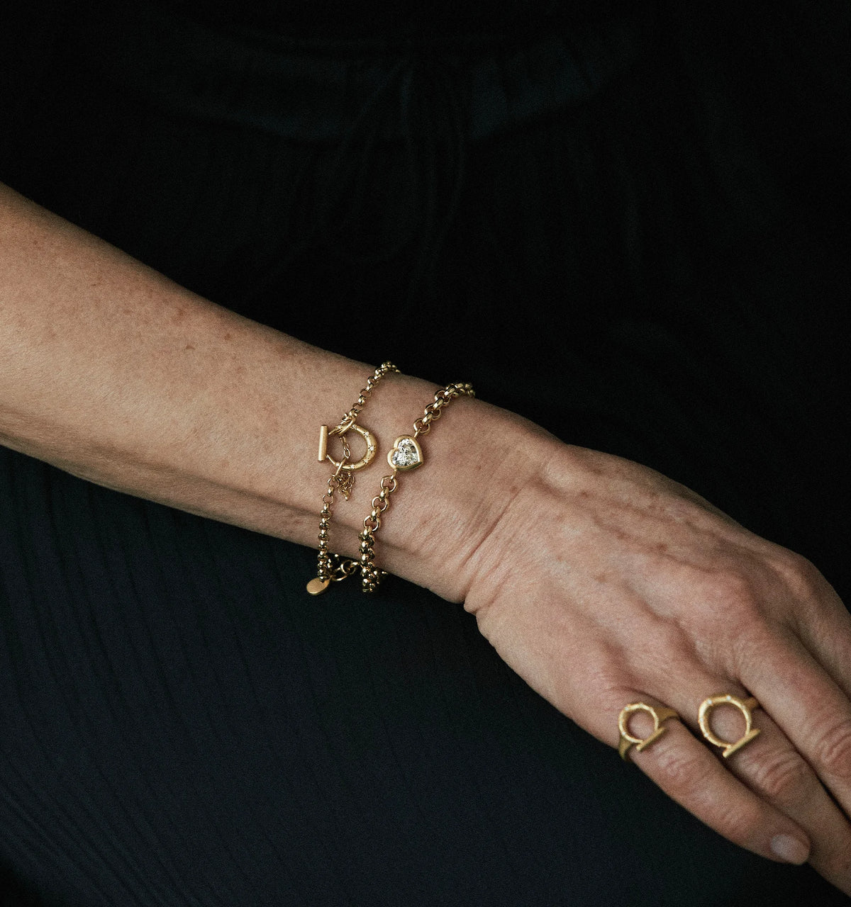 Woman wearing gold chain bracelets with heart charm and gold rings on her hand.
