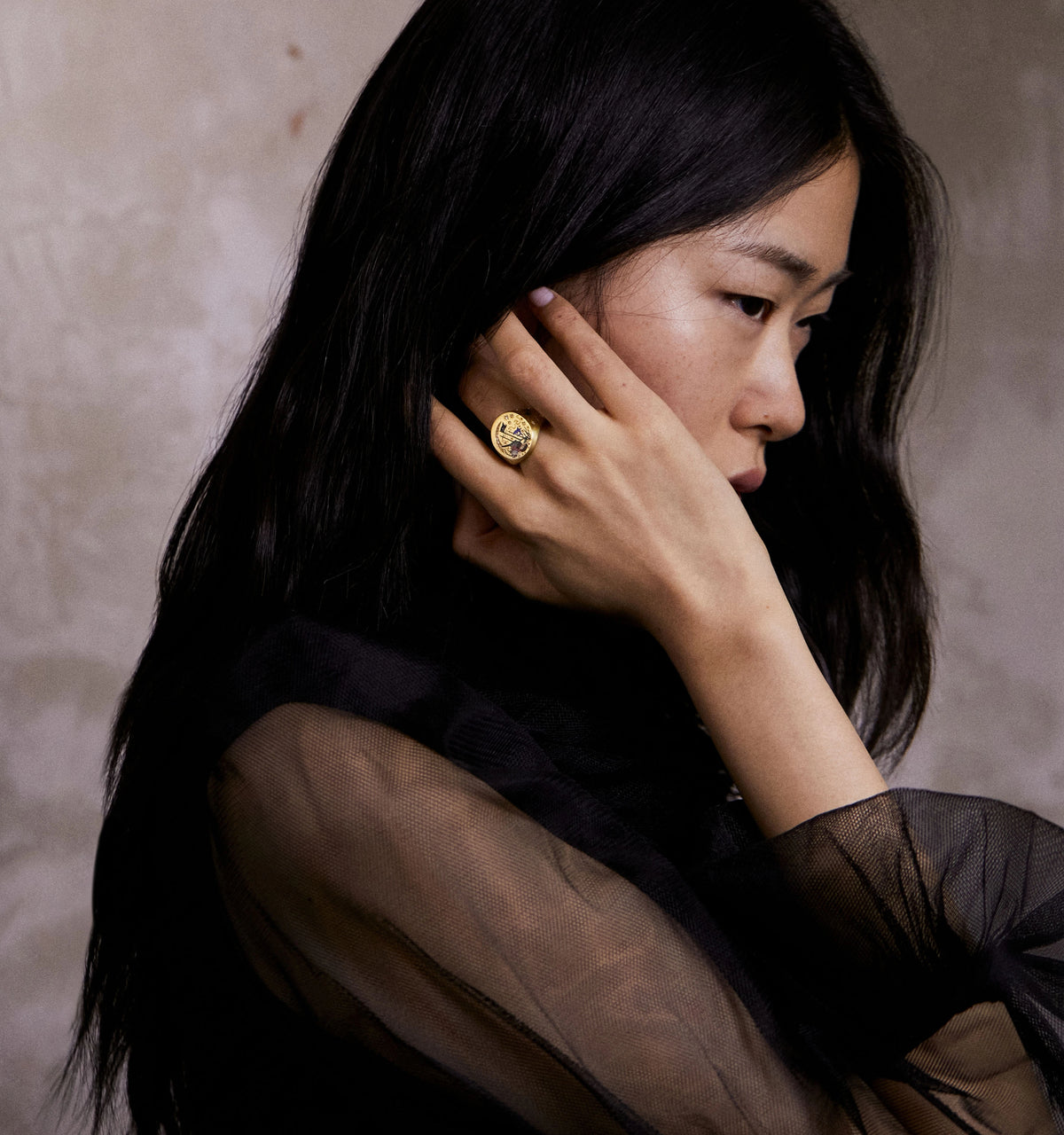 Woman wearing a gold ring with an intricate design and sheer black top, against a textured beige background.