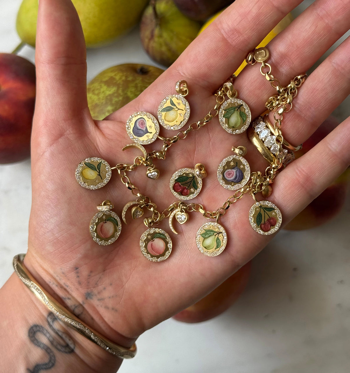 Gold charm bracelet featuring enamel fruit motifs, held in a hand with a gold bangle, surrounded by fresh fruits.