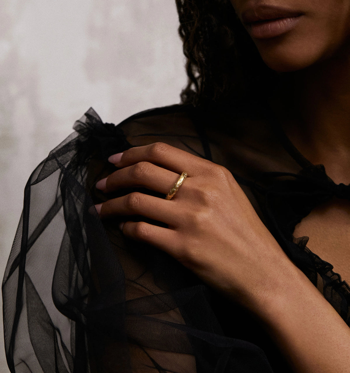 Woman wearing a textured gold ring, gently resting her hand on a sheer black fabric sleeve.