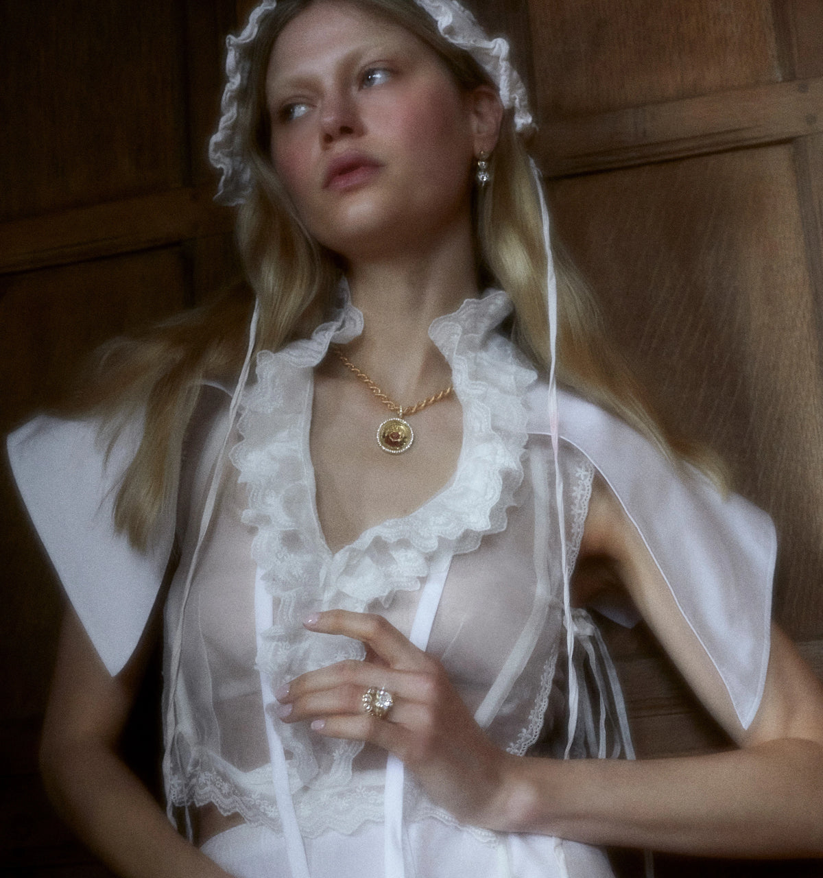 Woman wearing a gold necklace with a circular pendant and gold ring, styled in a sheer top with lace details.