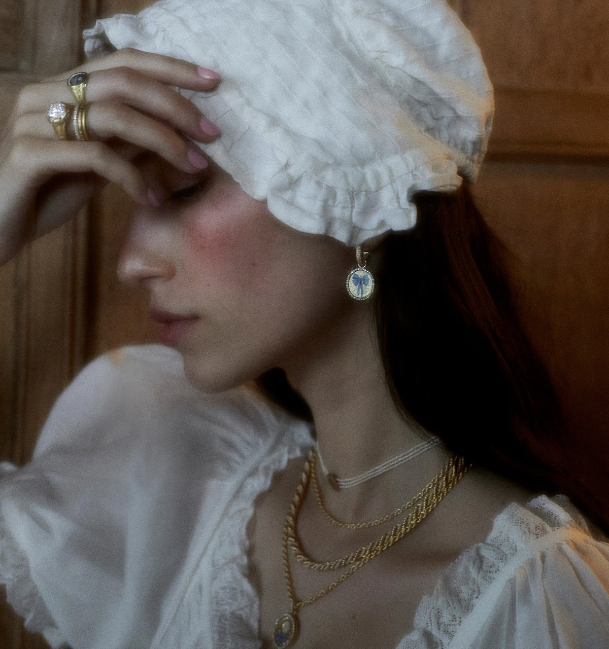 Woman wearing gold earrings and layered gold necklaces, styled with a white dress and cap, holding her forehead.