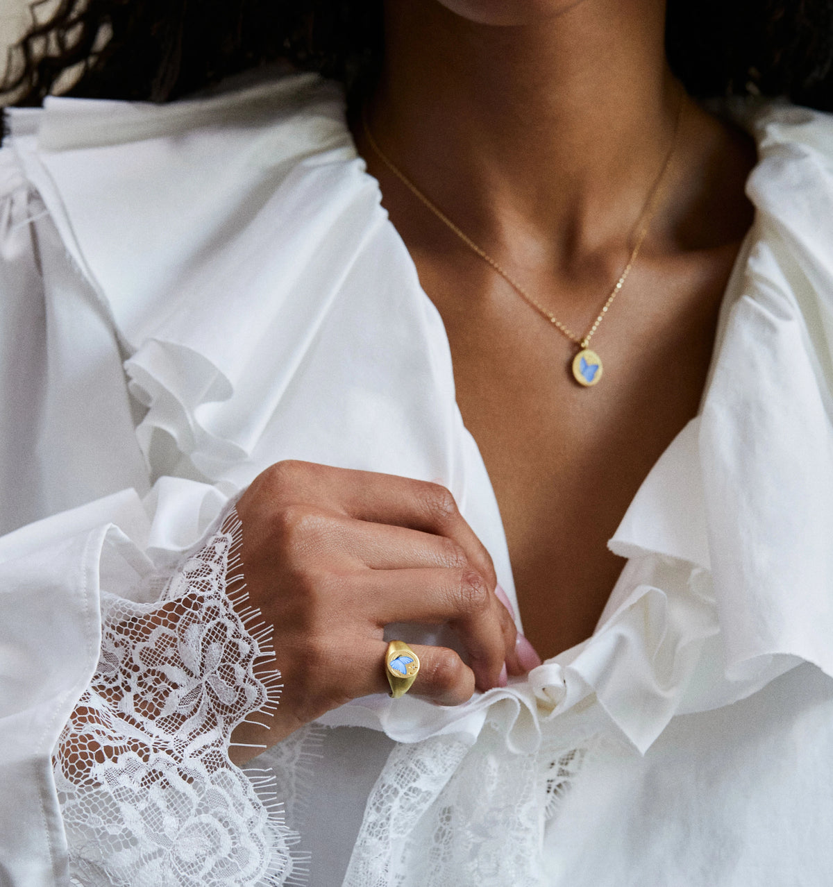 Woman wearing a gold necklace with a colourful pendant and a gold ring featuring a blue stone, dressed in a white blouse.