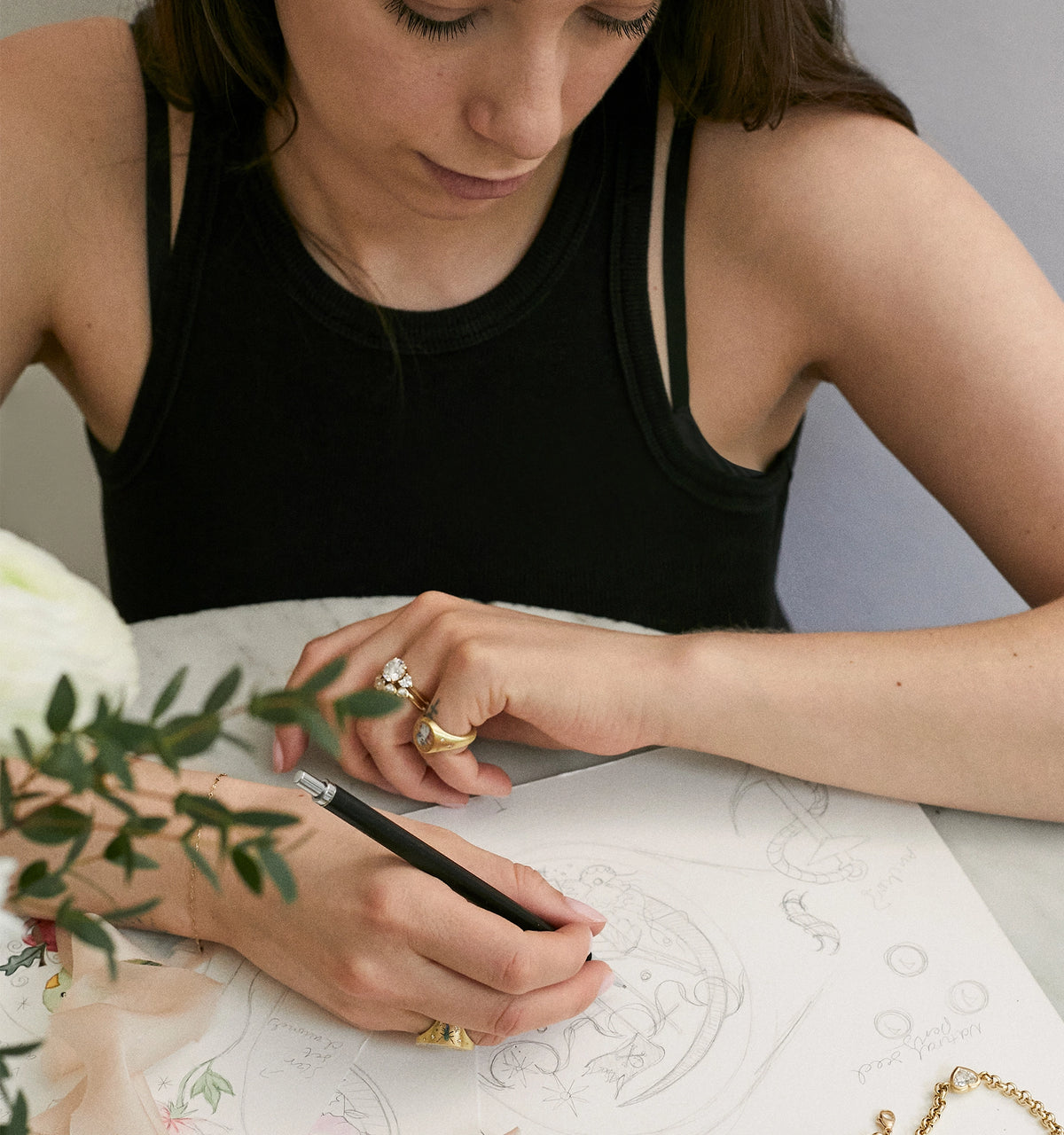 Woman wearing gold rings and a gold bracelet while sketching designs on paper at a marble table.