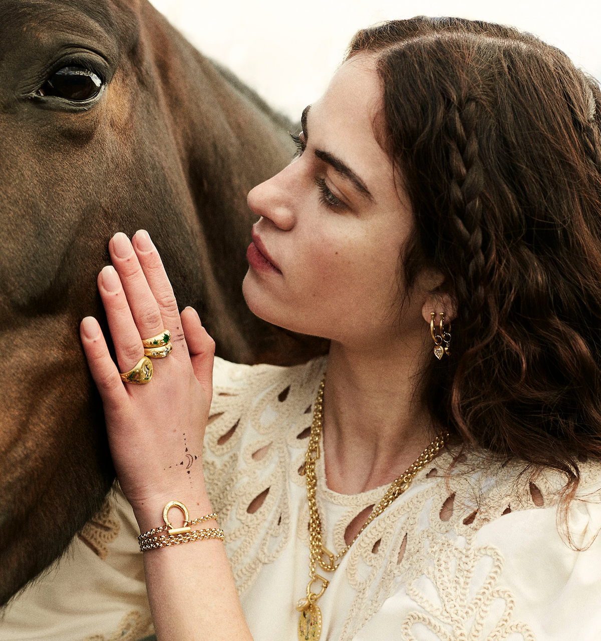 Woman wearing multiple gold rings and gold hoop earrings, gently touching a horse's face in a serene outdoor setting.