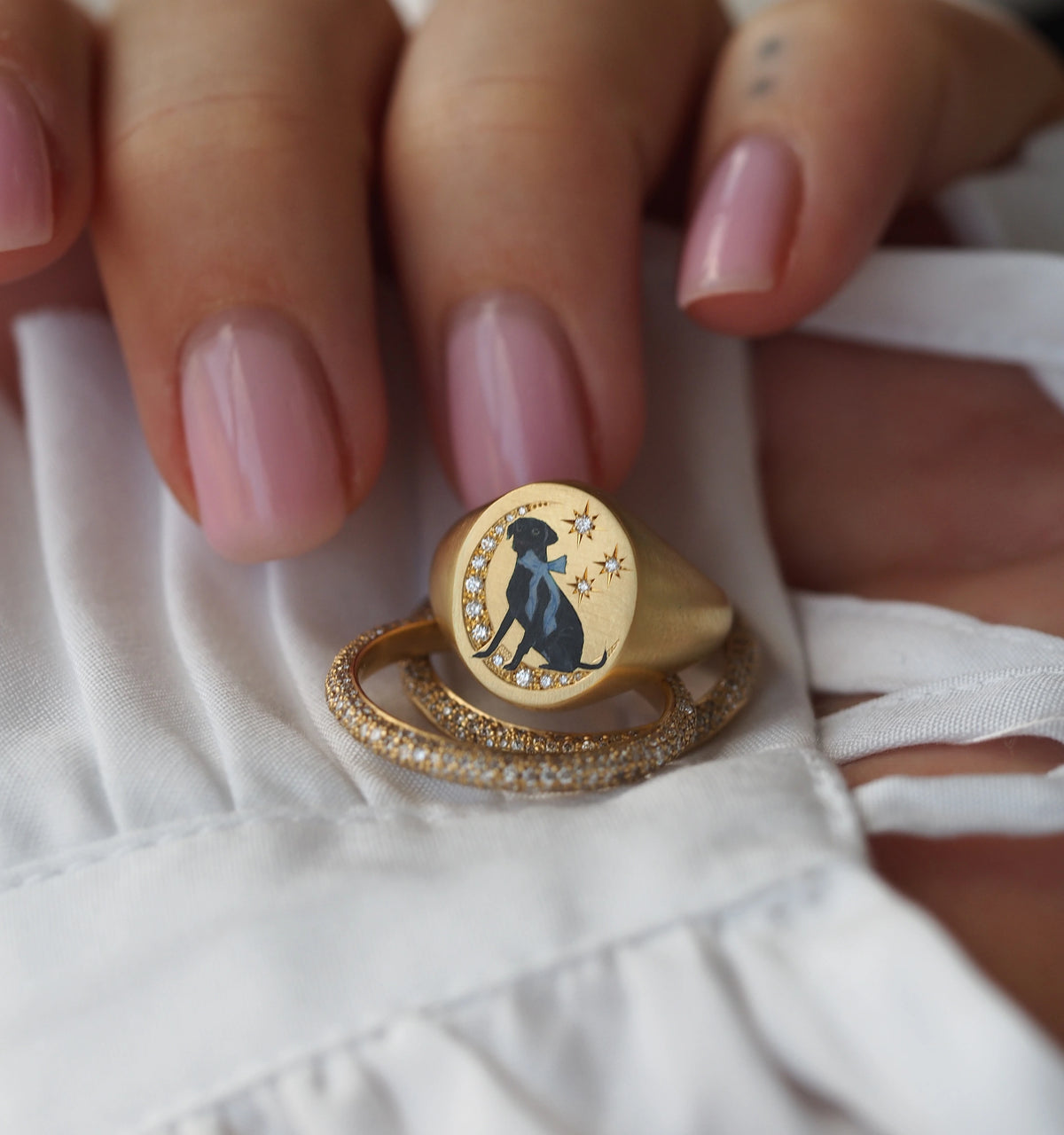 Woman wearing a gold ring with a black dog illustration and stars, resting on a white fabric surface.