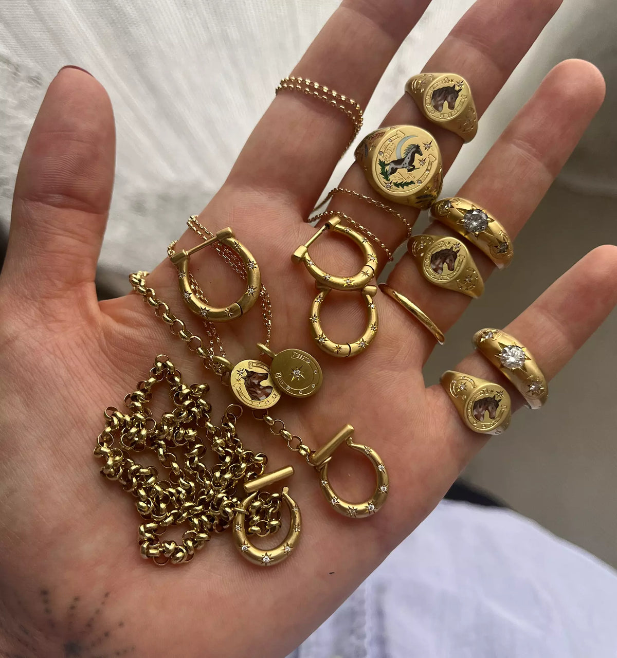 Hand holding multiple gold rings, gold hoop earrings, and a gold chain with a pendant, featuring intricate designs.