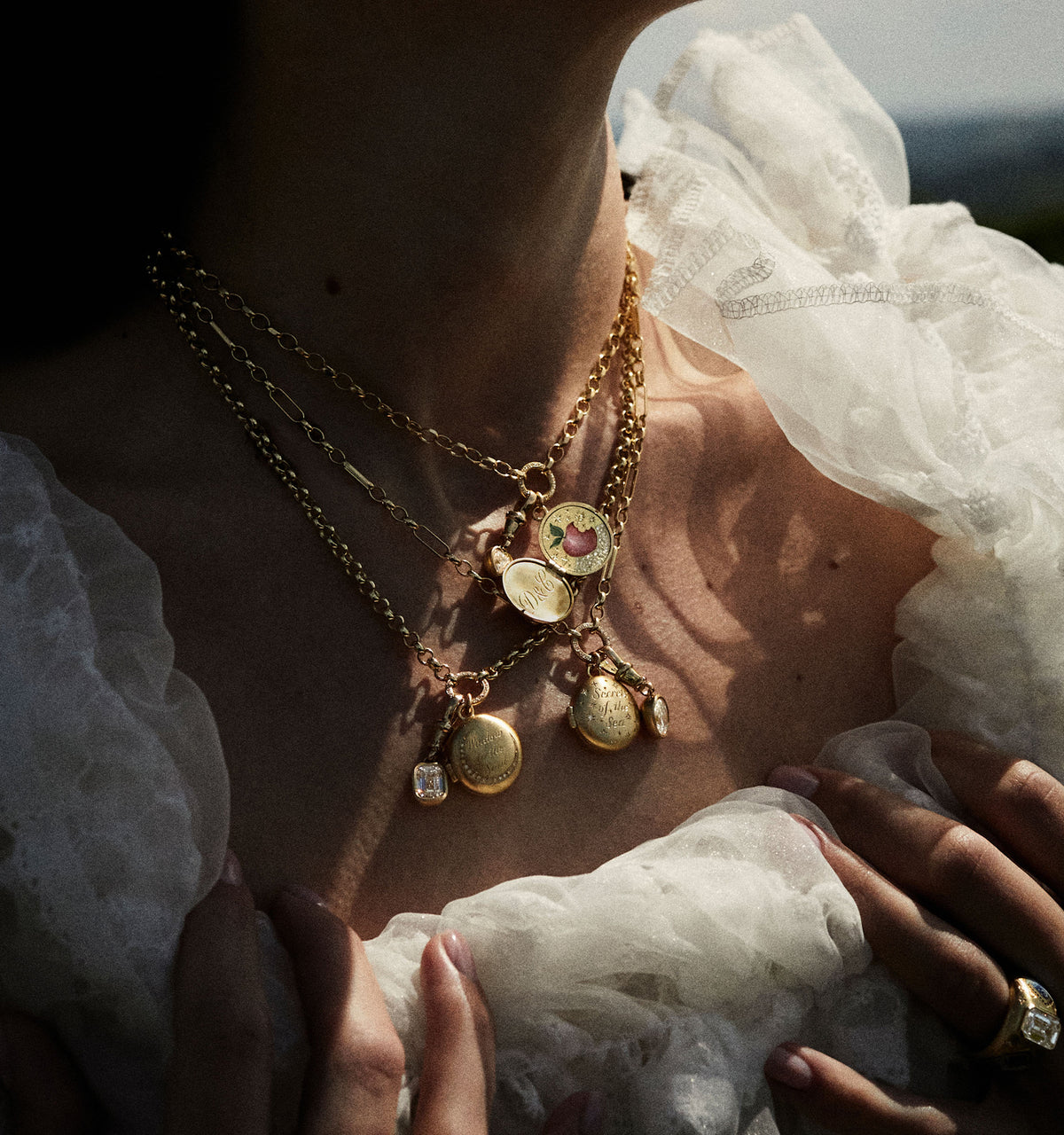 Woman wearing layered gold necklaces with engraved pendants and a gold ring, all set against a light, textured fabric.