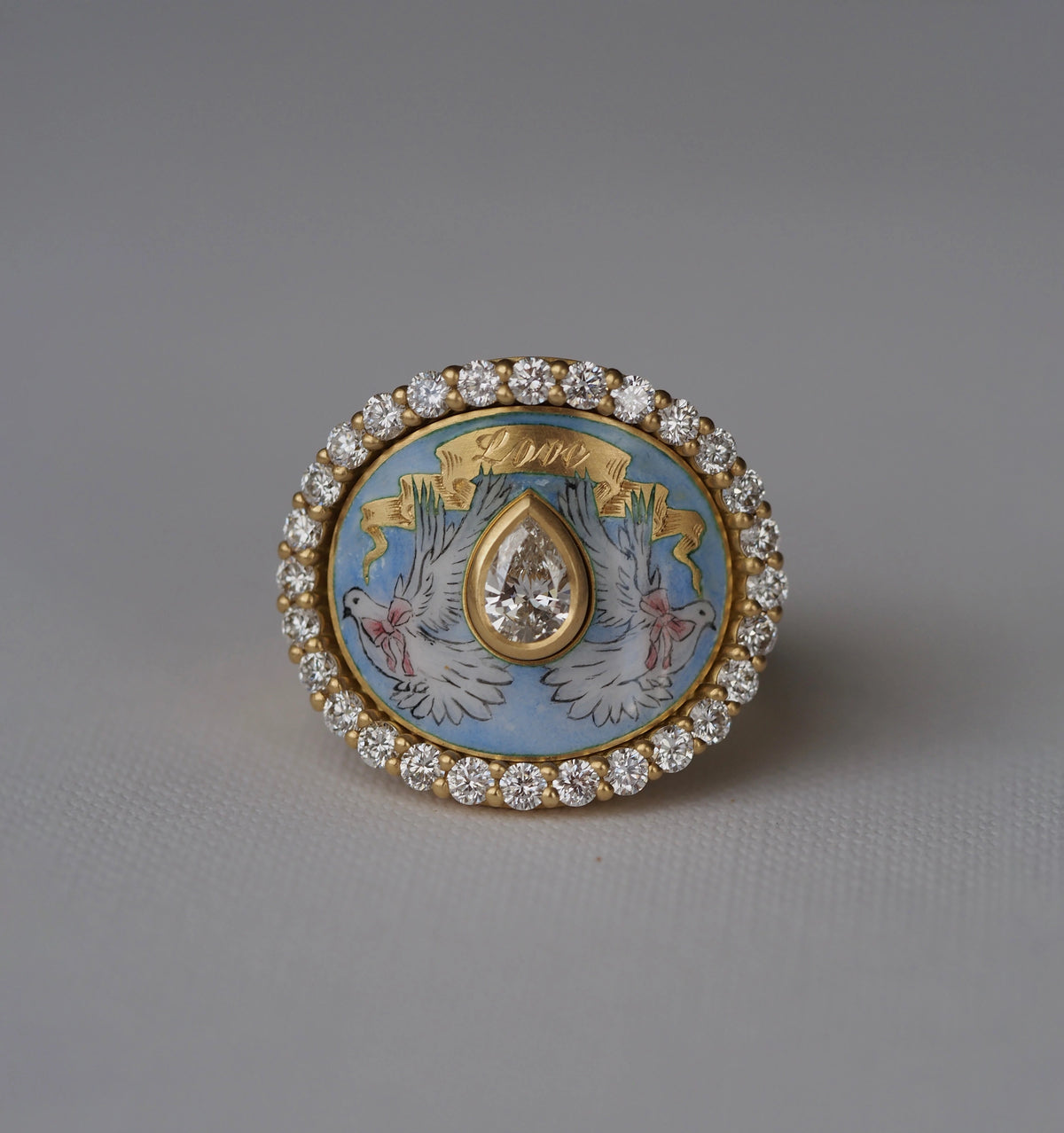 Gold ring featuring a blue enamel centre with doves and a drop-shaped diamond, surrounded by a halo of small diamonds.