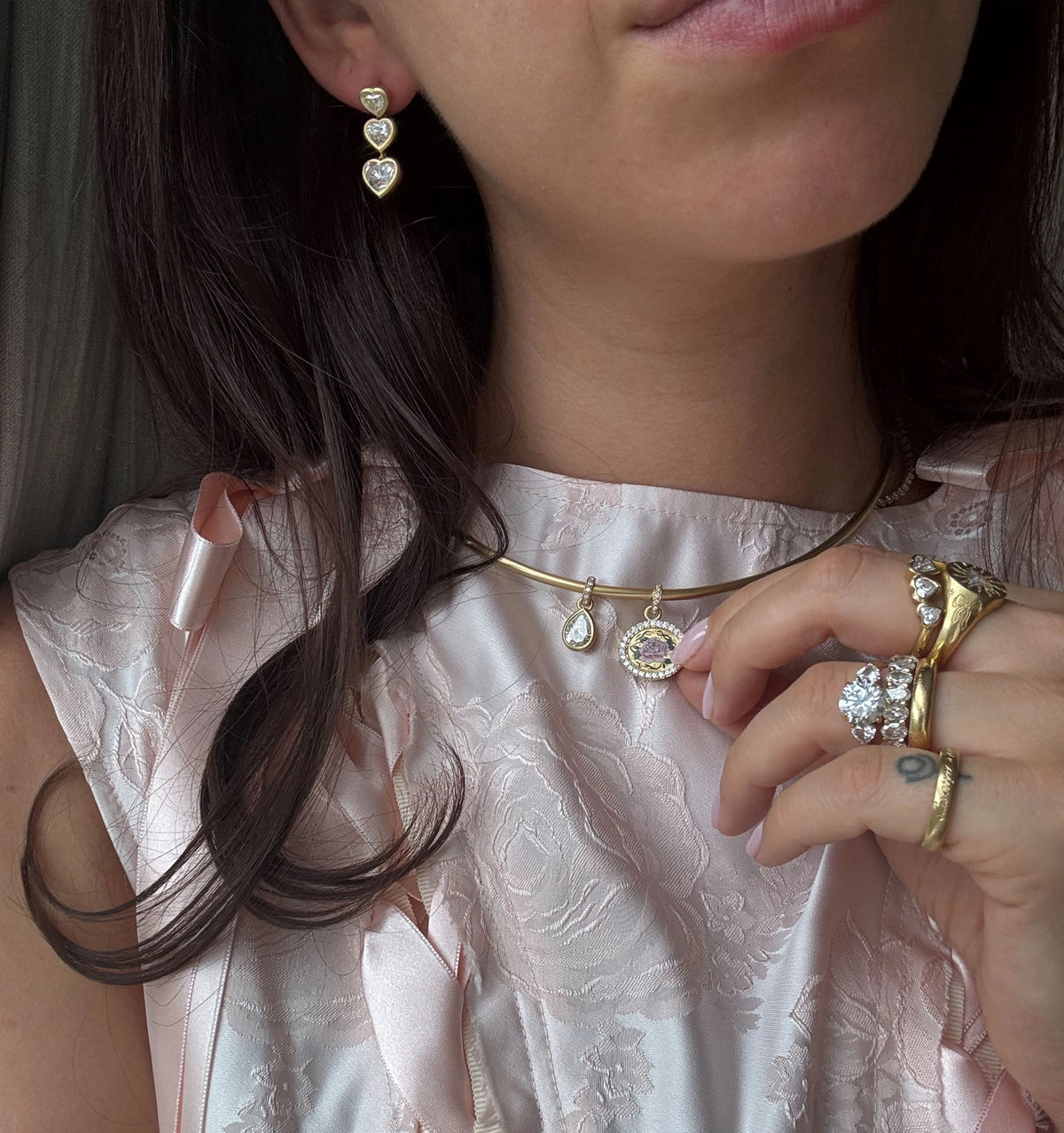 Woman wearing gold earrings, gold necklace with a teardrop pendant, and multiple gold rings, dressed in a pink floral top.
