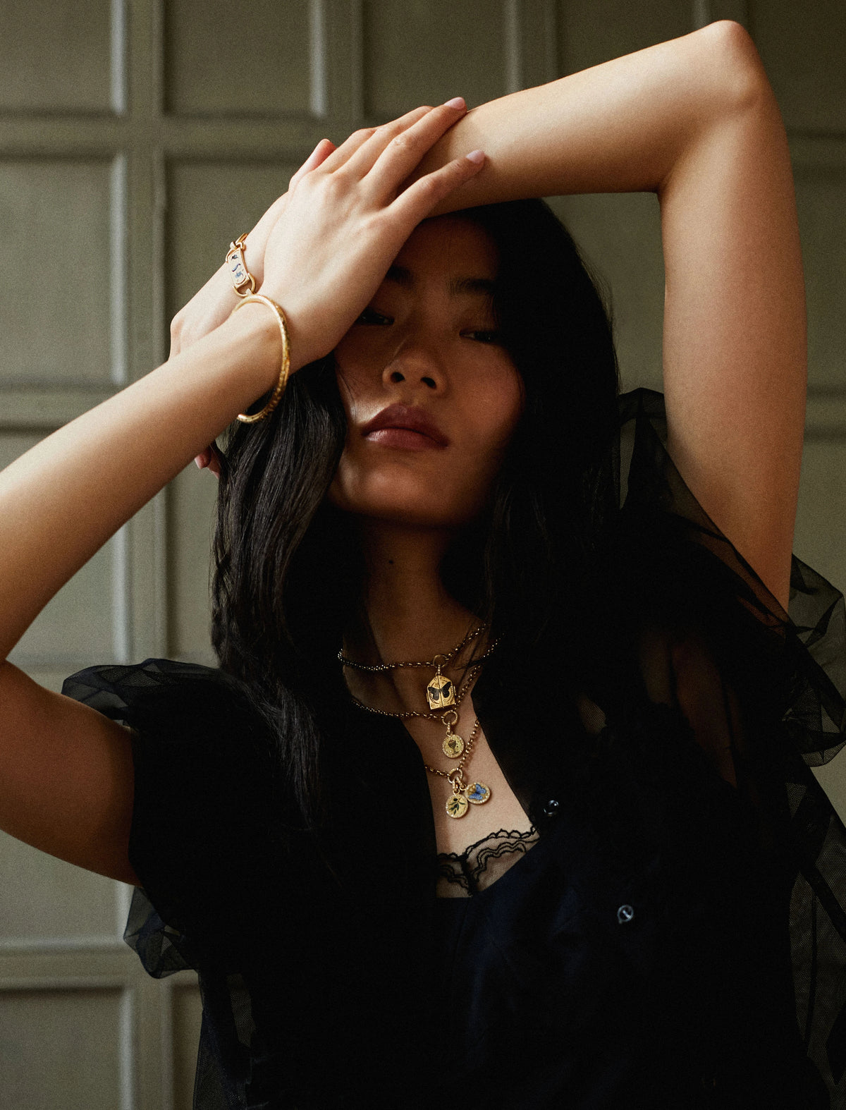 Woman wearing layered gold necklaces and a gold bracelet, with her arm raised above her head.