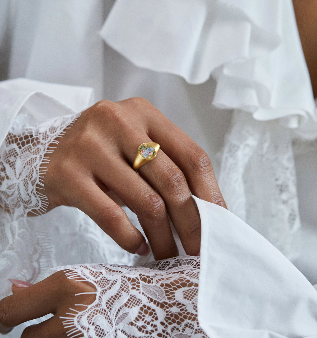 Woman wearing a gold ring featuring an enamel design with colourful floral motifs, resting on a lace-clad hand.