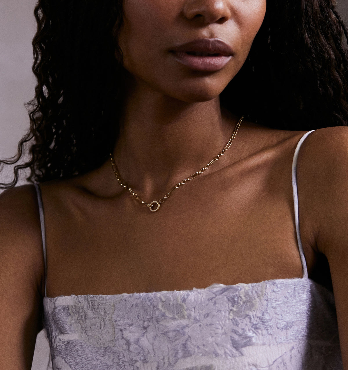 Woman wearing a gold chain necklace with a circular pendant, set against a neutral background.