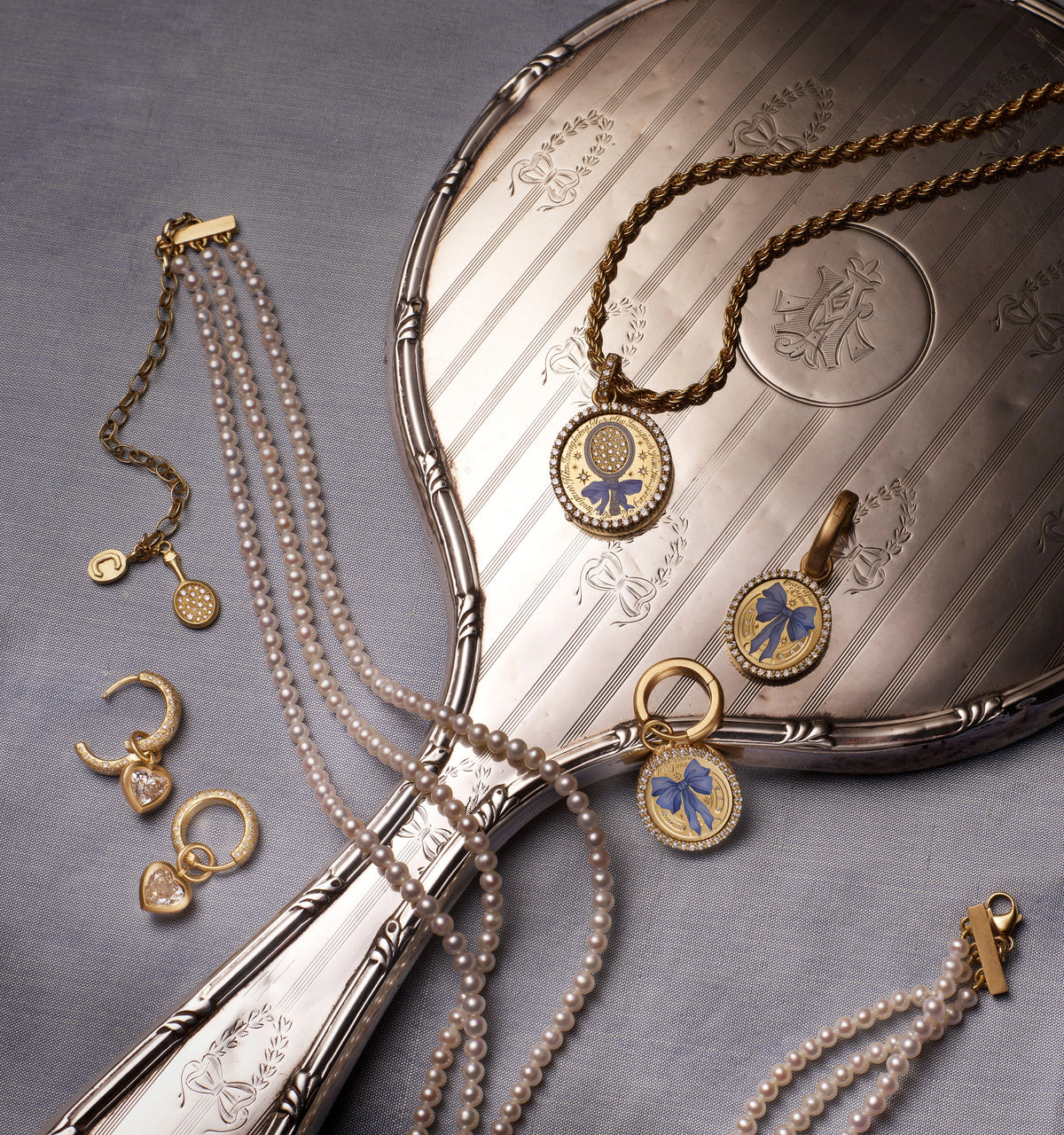 Gold pendants with blue enamel bow medallions and diamonds, pearl necklaces, and hoop earrings, on a silver hand mirror.