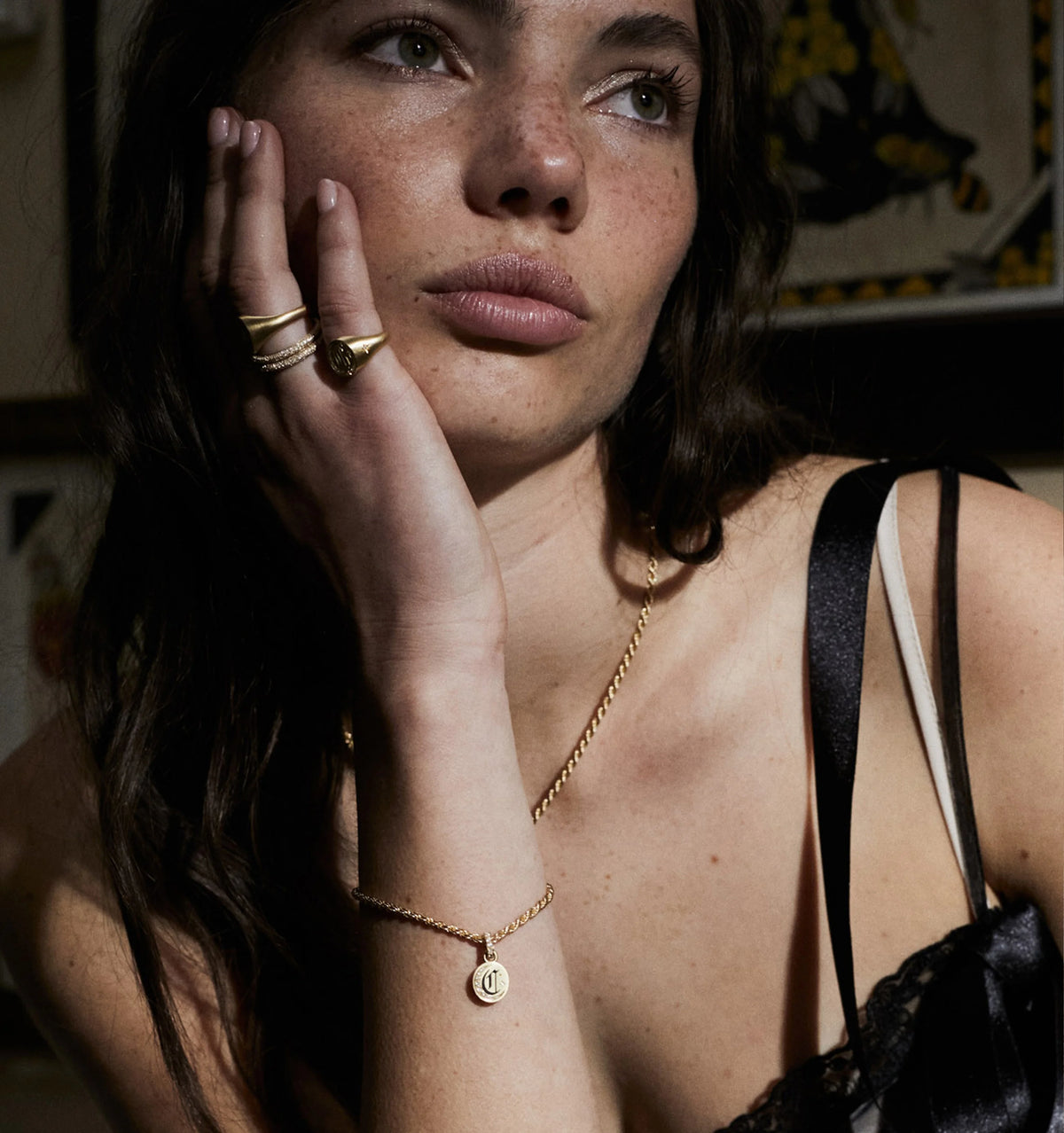 Woman wearing multiple gold rings and a gold bracelet with a charm, posing thoughtfully with her hand on her face.