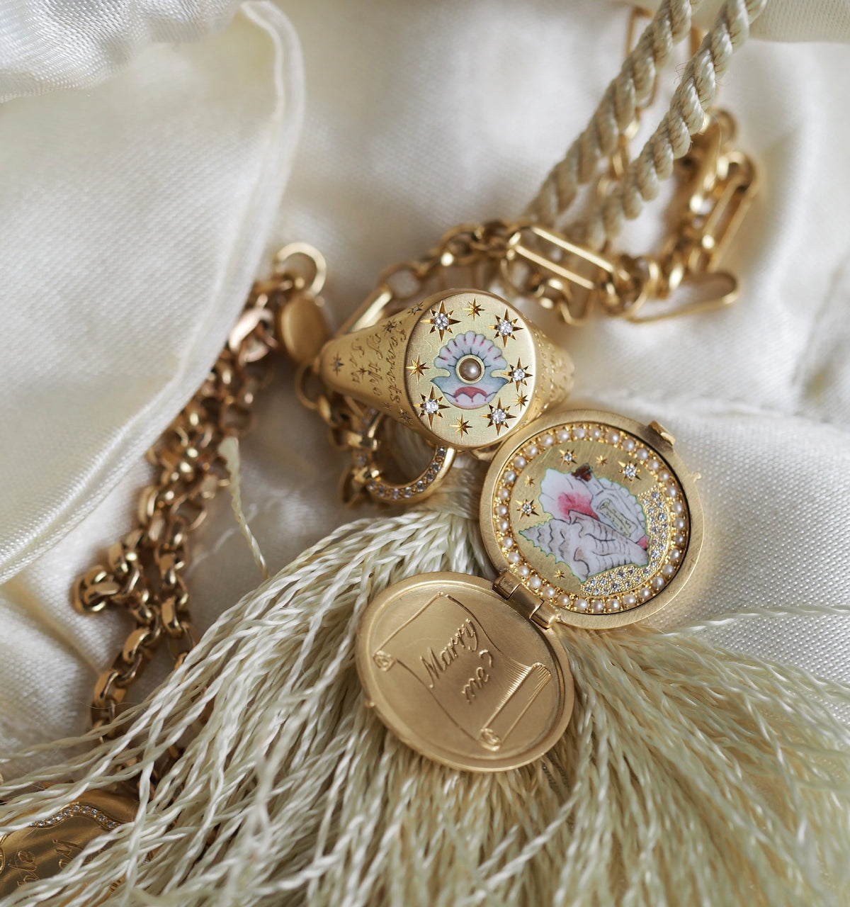 Gold ring with enamel floral design and a gold locket pendant featuring a painted scene, resting on a soft fabric background.