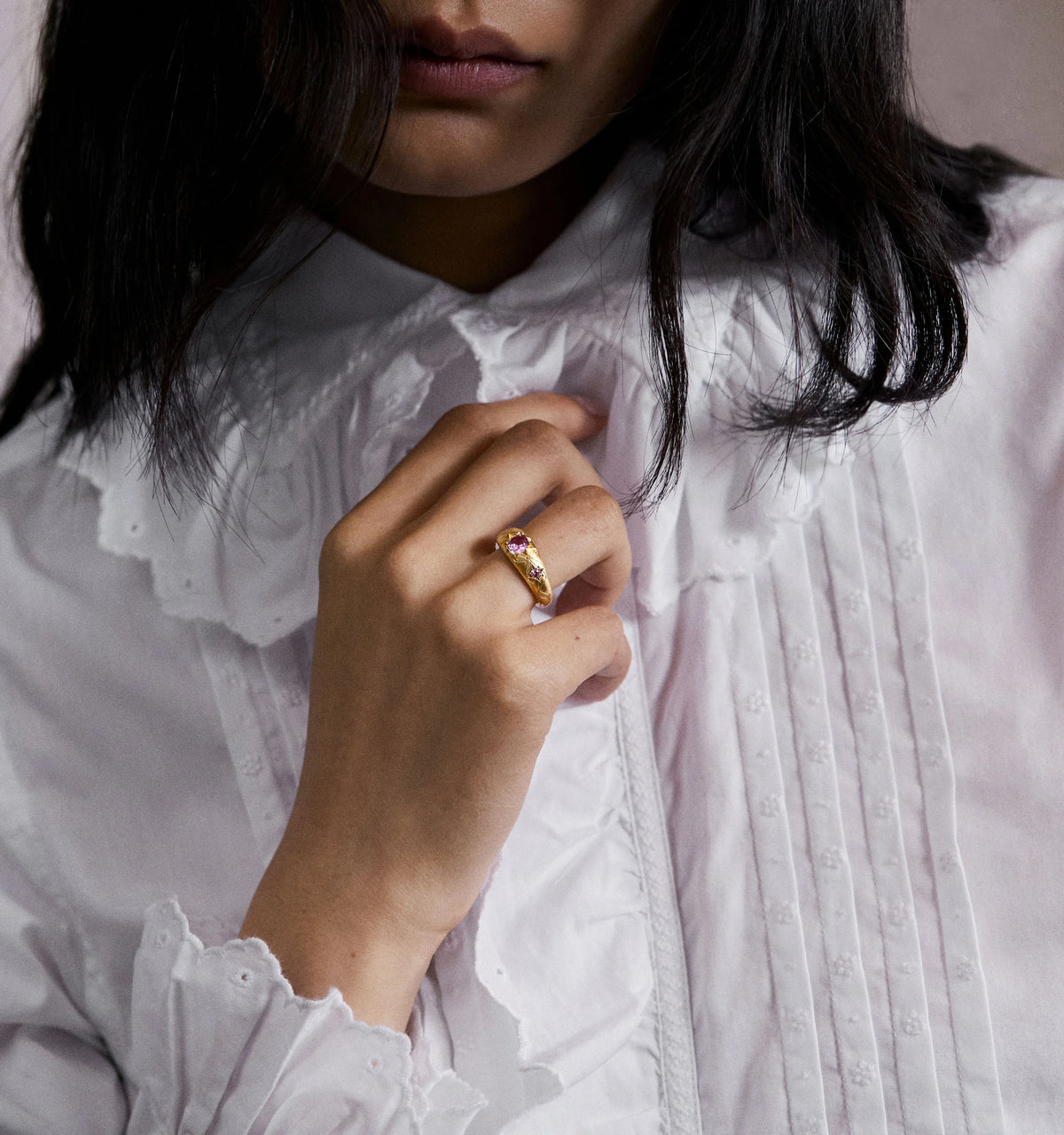 Woman wearing a gold ring set with pink sapphires, holding her hand gently against a textured white blouse.
