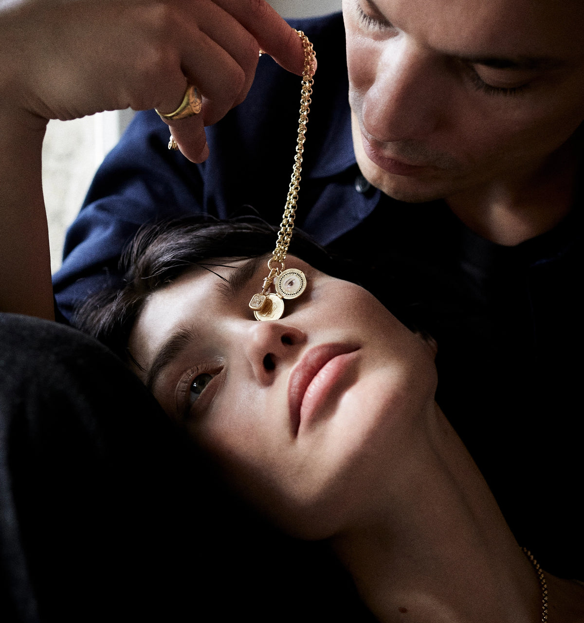 Woman wearing a gold necklace with charm pendants, resting against a man's arm in a softly lit setting.