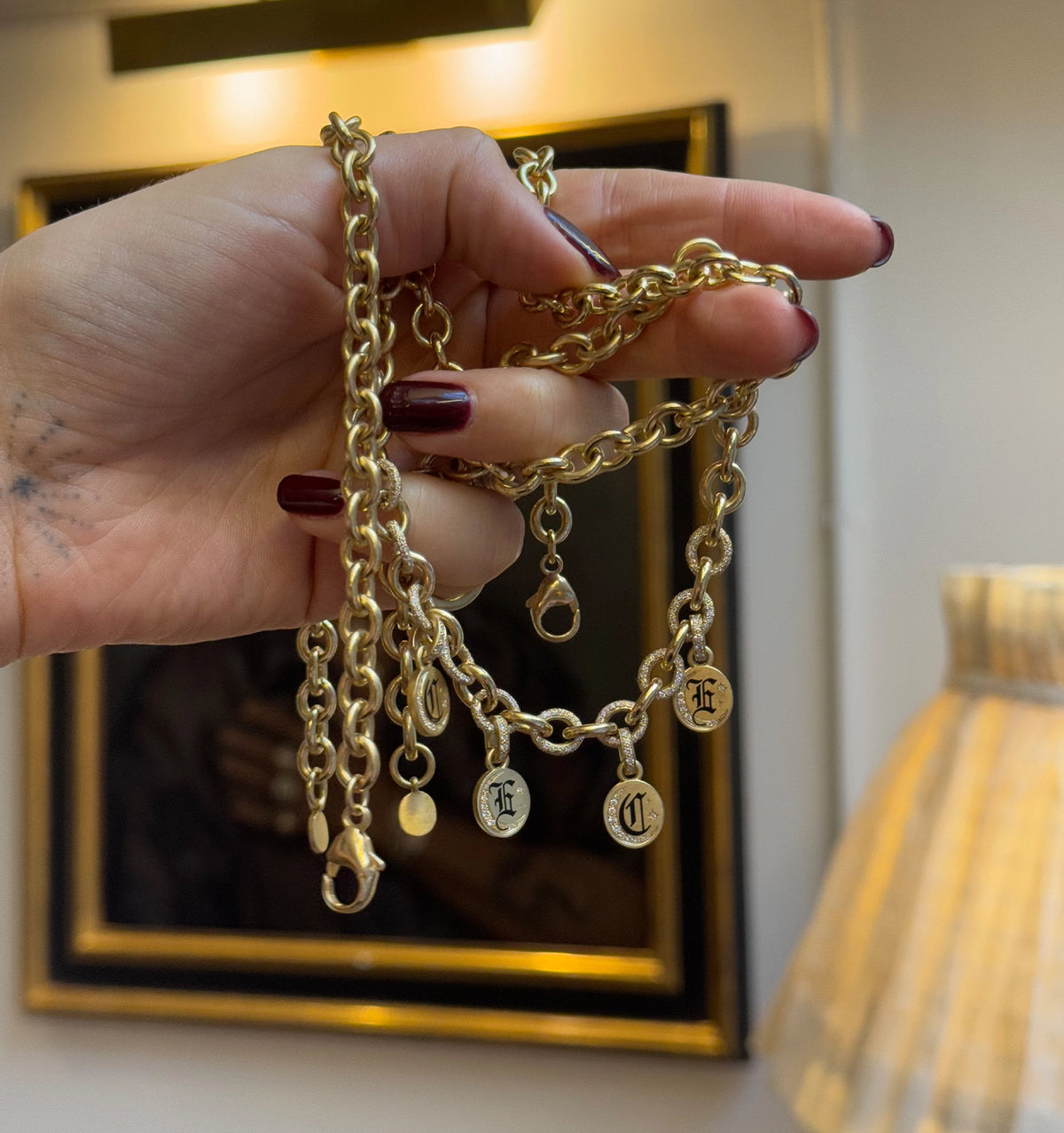 Gold chain necklace with engraved circular charms, held by a hand with long nails, set against a softly lit background.