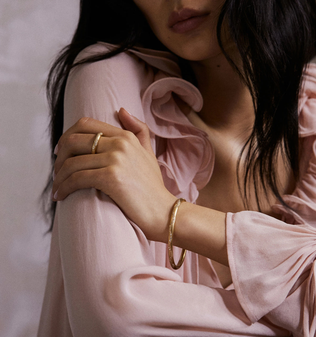 Woman wearing a gold bracelet and ring, with a soft pink blouse, resting her arms on a light-coloured surface.