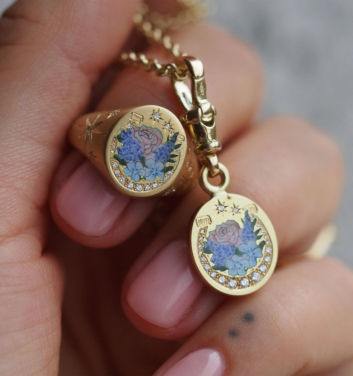 Gold ring and pendant featuring floral enamel design and diamond accents, held in a hand with pink nails.