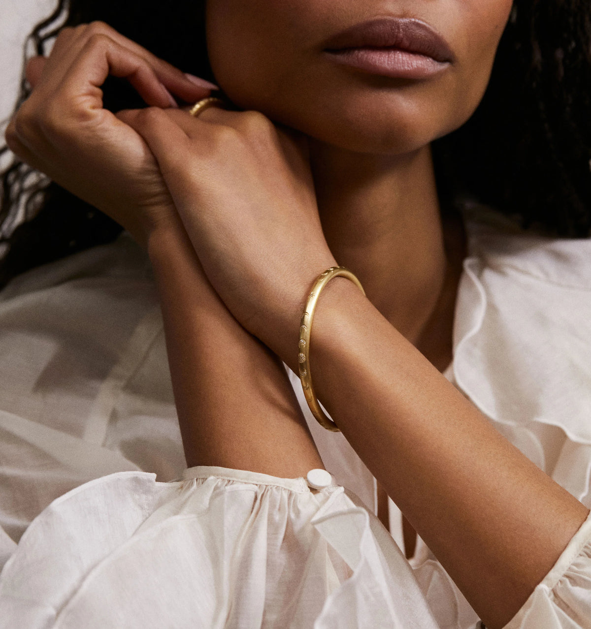 Woman wearing a gold bangle bracelet with a textured design, resting her arms casually on a light fabric.