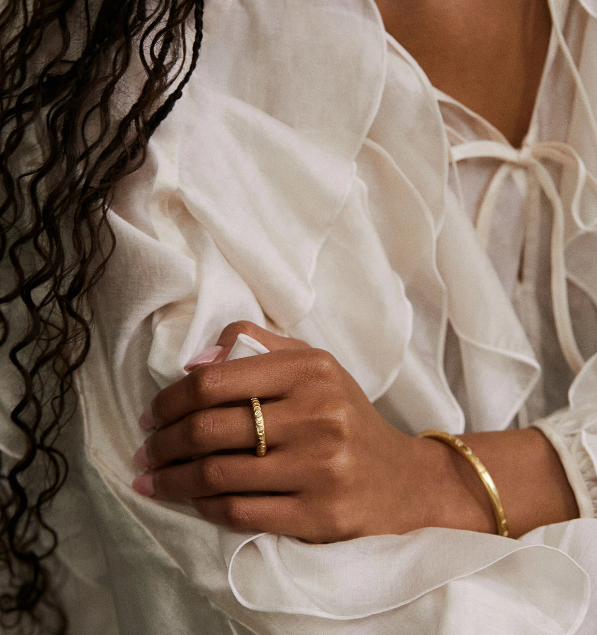 Woman wearing a gold textured ring and gold bangle, resting her hand on a white, flowing blouse.