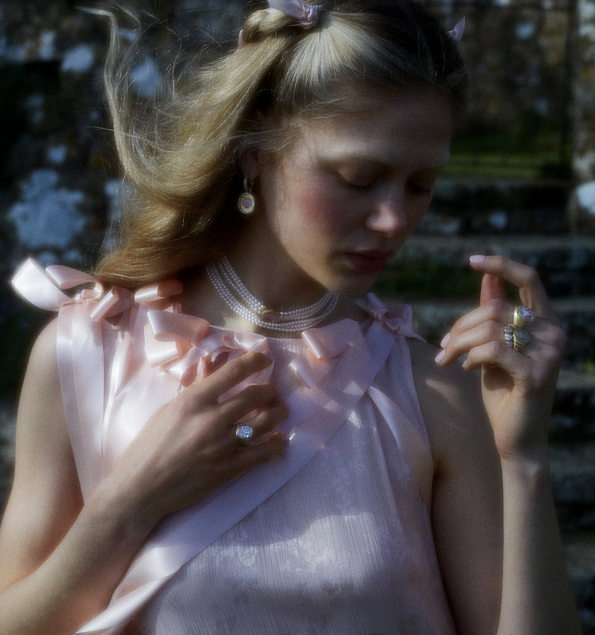 Woman wearing a gold ring and gold earrings, with a layered pearl necklace, dressed in a soft pink outfit.