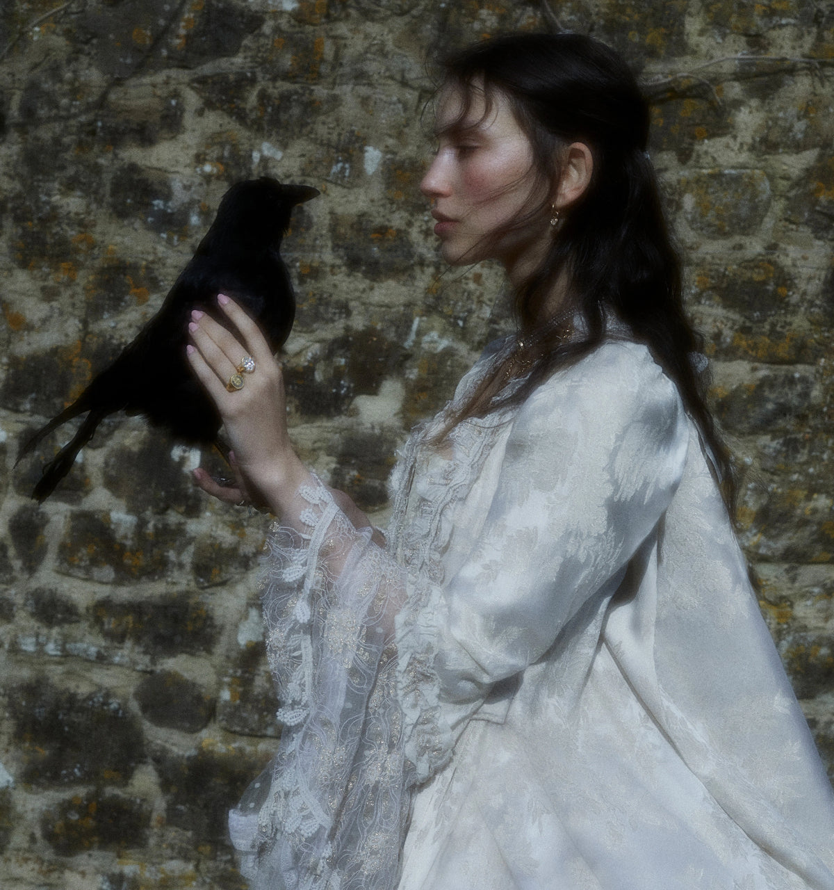 Woman wearing gold ring and gold earrings, holding a black crow against a textured stone wall.