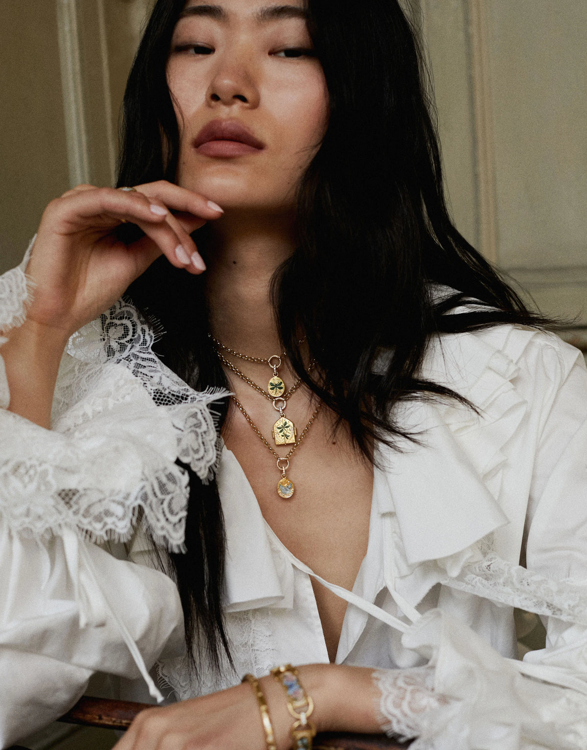 Woman wearing layered gold necklaces with coloured enamel motifs and a gold bracelet, dressed in a white ruffled blouse.
