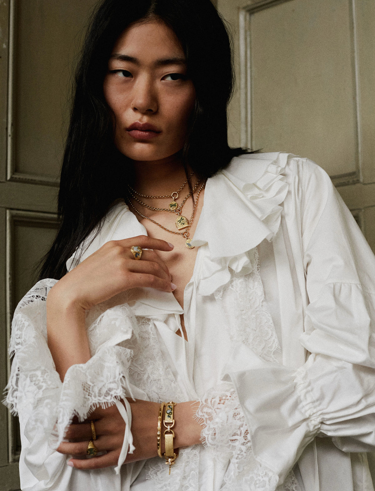 Woman wearing layered gold necklaces, gold ring, and gold bracelet, styled with a white ruffled blouse.