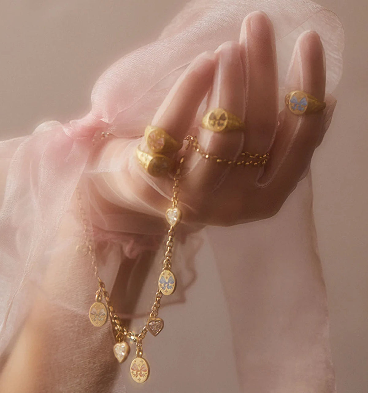 Woman wearing multiple gold rings and a gold necklace with decorative pendants, draped in soft pink fabric.