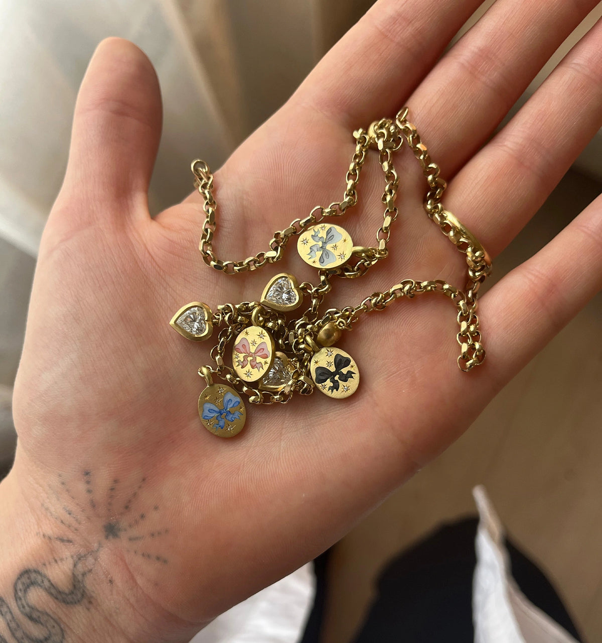 Gold necklace with bow enamel charms in blue, pink, and black, and heart-shaped diamond charms, held in a hand.