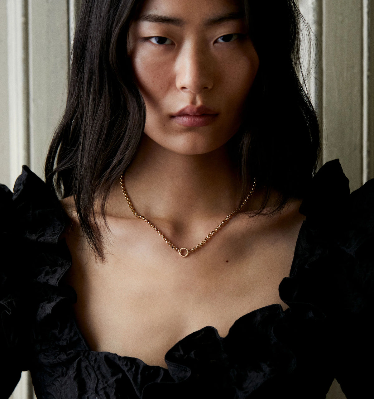 Woman wearing a gold necklace featuring a circular pendant, styled with a ruffled black top.