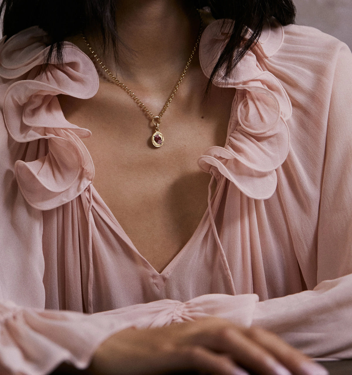 Woman wearing a gold necklace with a ruby pendant and a soft pink blouse with ruffled details.