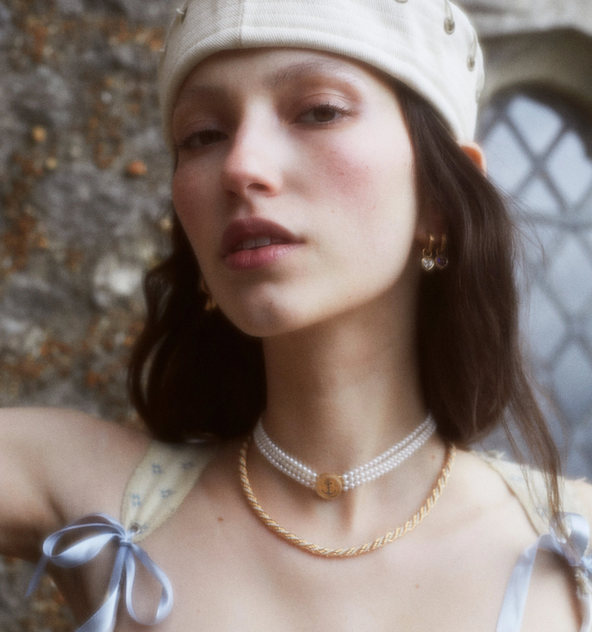 Woman wearing gold hoop earrings and layered gold necklaces with a gold pendant, styled with a white top and a cap.