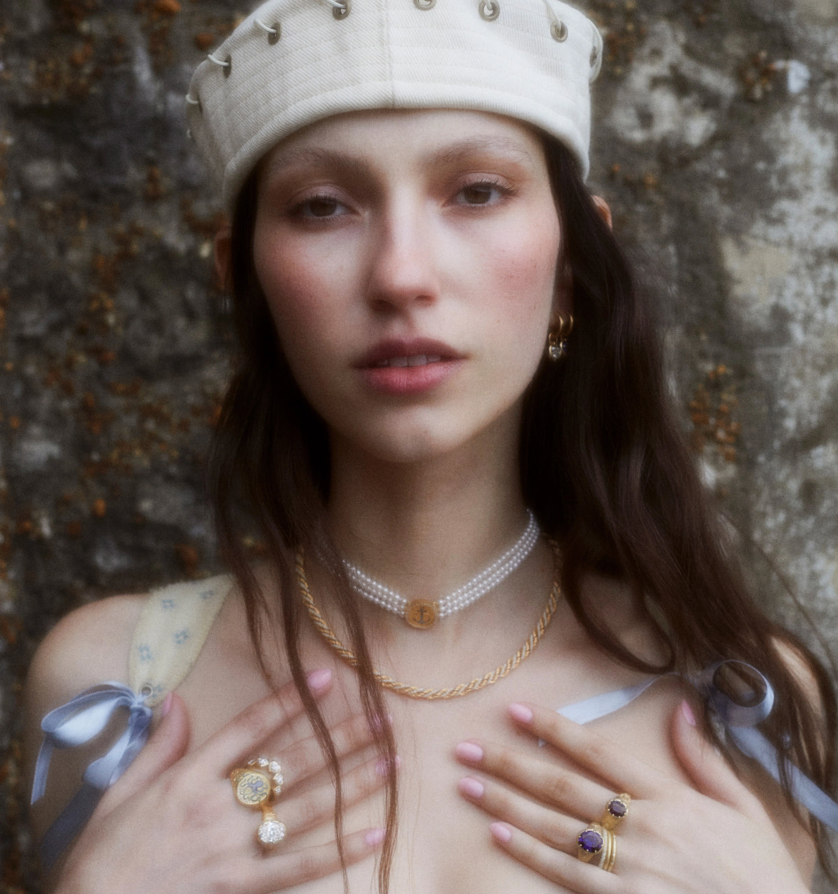 Woman wearing gold necklaces and rings, including a blue sapphire ring, against a textured stone background.