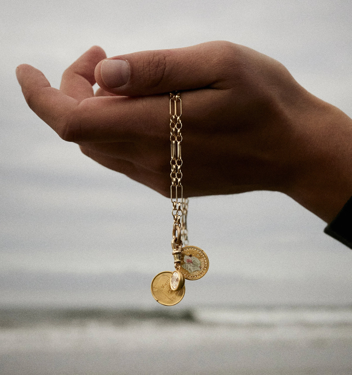 Gold chain bracelet with circular pendants held by a hand against a beach background.