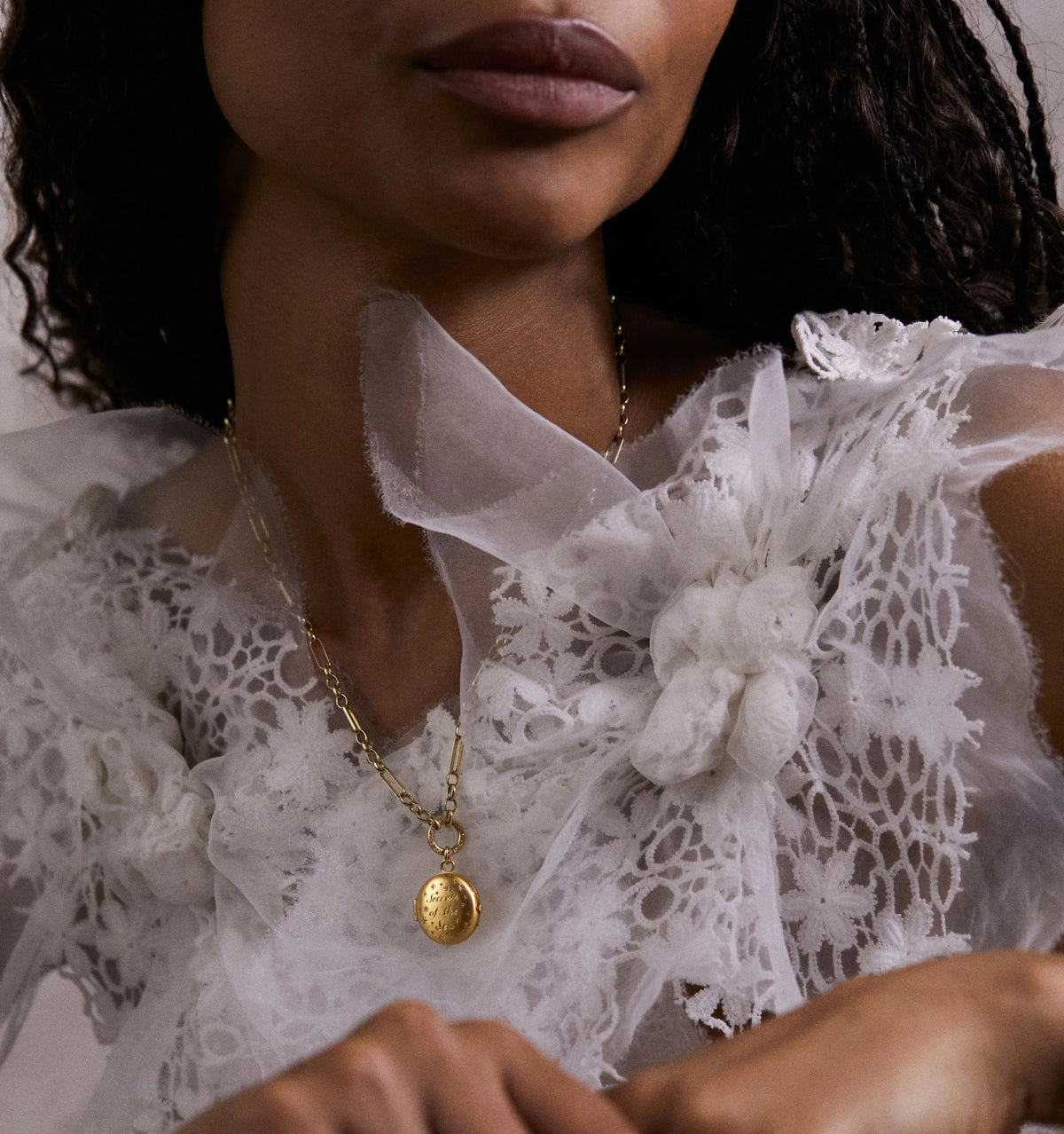 Woman wearing a gold pendant necklace with engraved details, paired with a delicate white lace top.