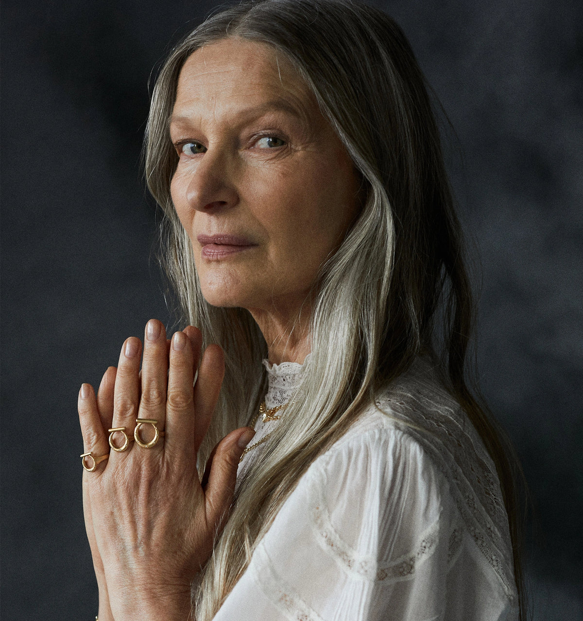 Woman wearing three gold rings with a simple design, hands clasped together against a dark textured background.