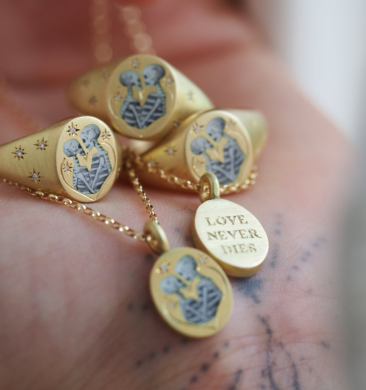 Gold rings and pendant featuring engraved skeleton couple design and "LOVE NEVER DIES" inscription on a hand.