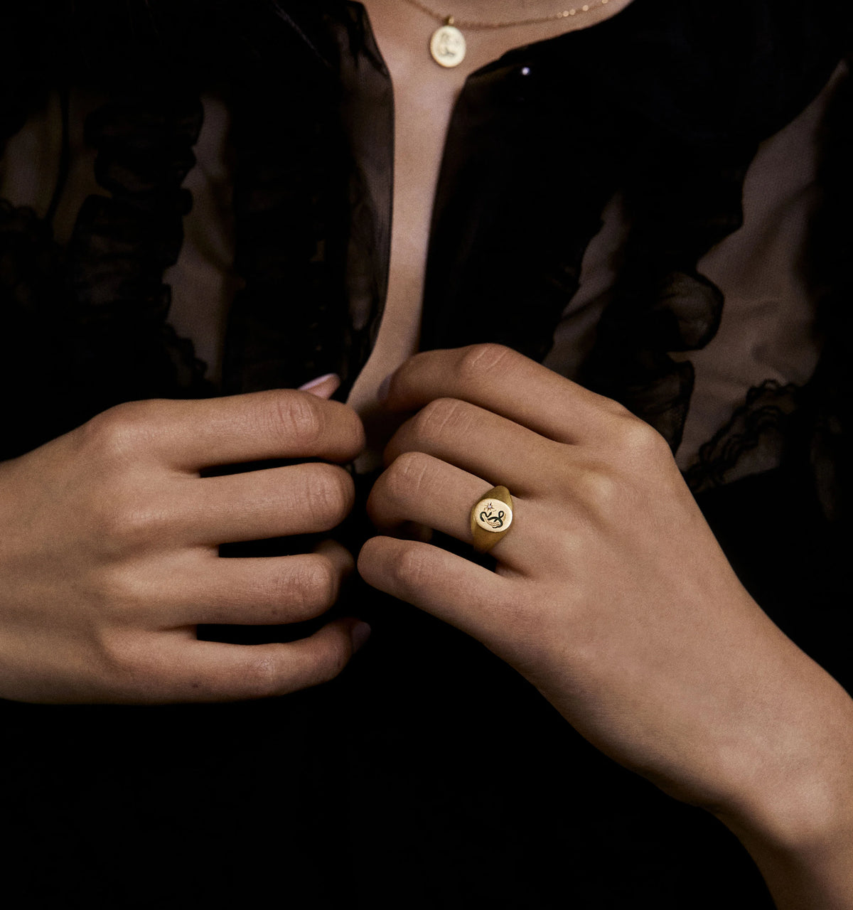 Woman wearing a gold ring with a heart design, holding her hands together in front of a black ruffled blouse.