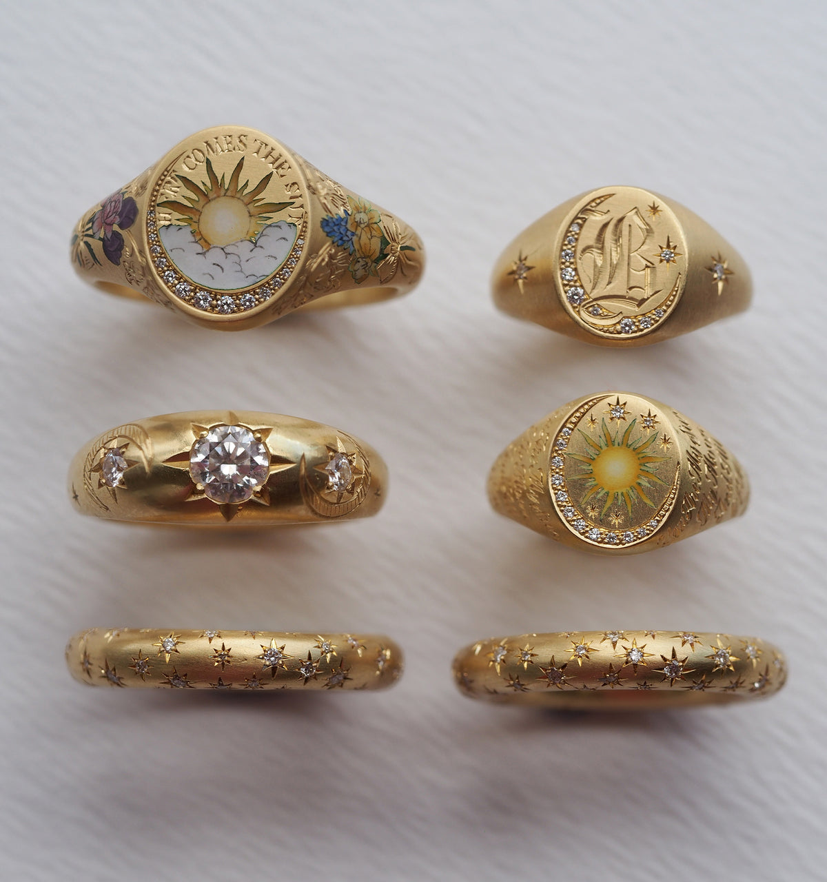 Gold rings featuring engraved designs, colourful motifs, and a central diamond stone, arranged artistically on a textured surface.