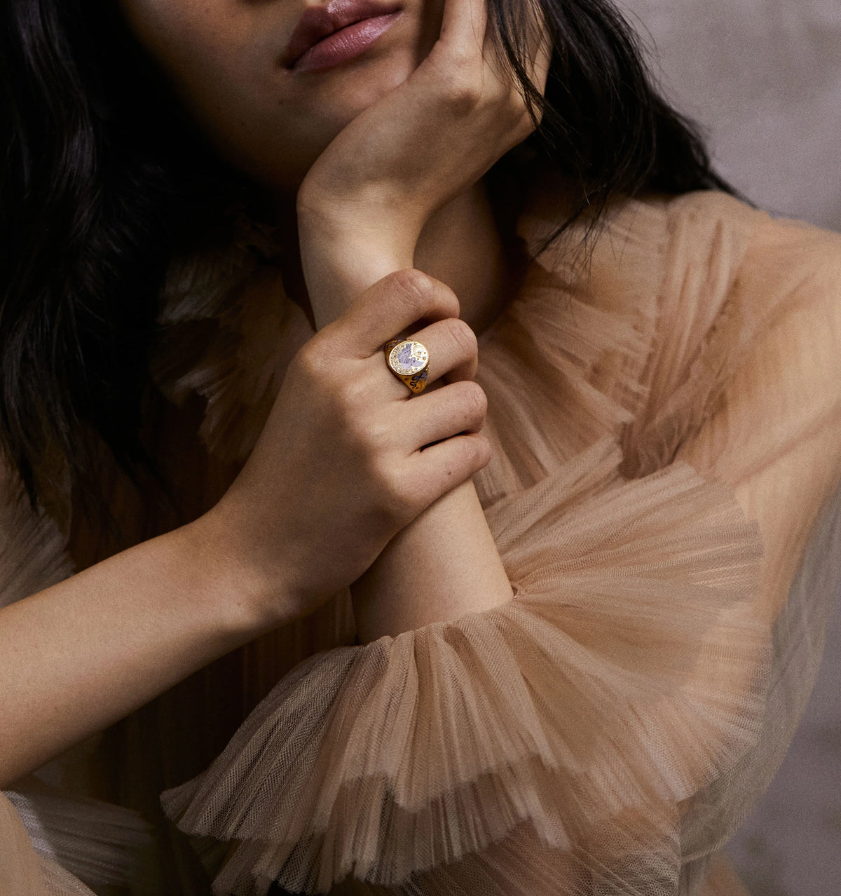 Woman wearing a gold ring with a heart motif, resting her chin on her hand, in a softly lit setting.